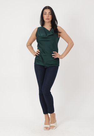 Donna con capelli scuri che indossa una canotta verde scuro senza maniche, jeans skinny scuri e sandali bianchi con tacco, in piedi con le mani sui fianchi.