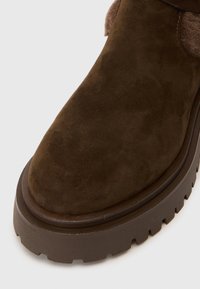 Bota de ante marrón con suela de goma gruesa y estriada, con forro de felpa visible en la abertura del tobillo.