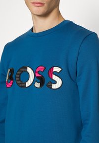 Blå sweatshirt avtexturerat tyg med rund halsringning. Har en iögonfallande "BOSS"-logotyp i flera färger: rosa, svart, vit och med texturerade detaljer.