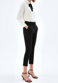 Vrouw in een witte blouse met zwarte strik, zwarte slim-fit cropped broeken en zwarte enkellaarsjes met bandje, staand met handen in haar zakken.