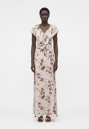 Γυναίκα φορώντας μακρύ ρόζ φόρεμα με λουλουδάτο σχέδιο, βολάν στο ντεκολτέ και κορδέλα στη μέση, στέκεται μπροστά σε ένα απλό λευκό φόντο.