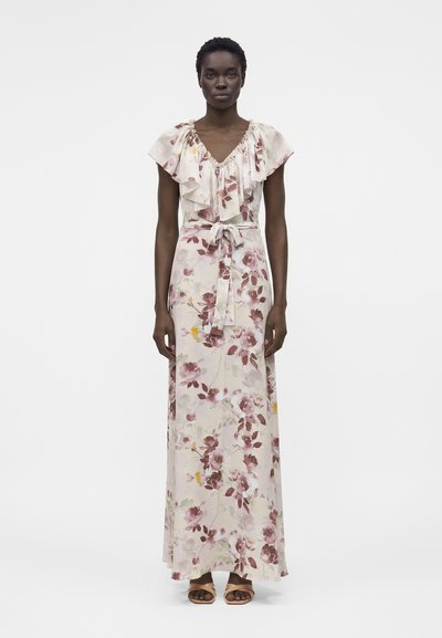 Mujer vistiendo un vestido largo hasta el suelo de color rosa claro con estampado floral, con escote fruncido y lazo en la cintura, de pie frente a un fondo blanco liso.