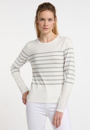 Jonge vrouw met licht haar draagt een wit shirt met lange mouwen met horizontale grijze strepen en een witte broek, staand met één hand op de heup.