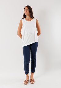 Top bianco senza maniche con orlo asimmetrico, abbinato a pantaloni aderenti blu navy e sandali marroni con zeppa, su uno sfondo neutro.