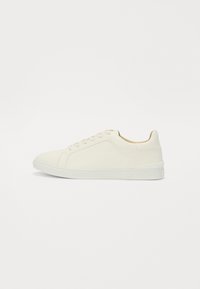 Sneaker blanche avec une tige en cuir lisse et une semelle en caoutchouc texturé, dotée de lacets plats et de détails de couture latérale minimalistes.