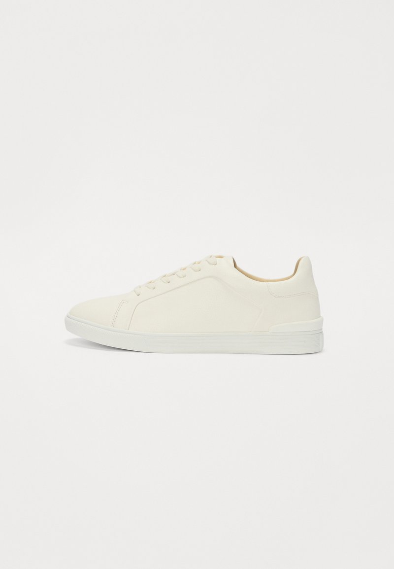 Sneaker blanche avec une tige en cuir lisse et une semelle en caoutchouc texturé, dotée de lacets plats et de détails de couture latérale minimalistes.