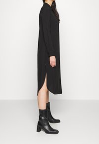 Robe chemise noire à boutonnage avec une coupe droite, fentes latérales et manches longues, associée à des bottines noires à semelle épaisse. Texture lisse.