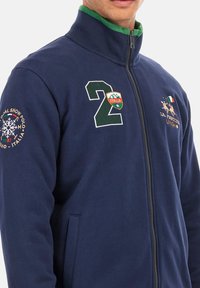Marinblå zip-up jacka i mjukt tyg, med gröna krageaccenter. Broderad siffra "2" och logotypmärken ger en extra detalj.
