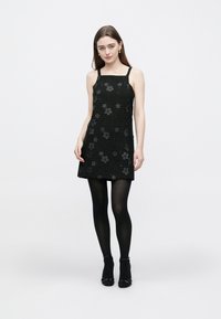 Juoda be rankovių suknelė su tekstūruota medžiaga, puošta gėlių aplikacijomis, virš kelių ilgio, derinama su juodomis pėdkelnėmis ir aukštakulniais batais.