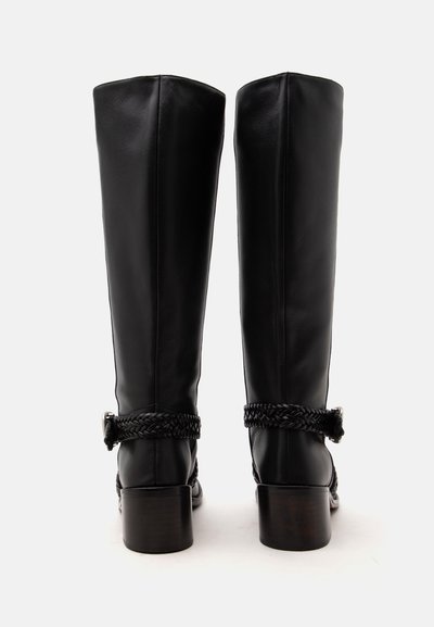 Paire de bottes en cuir noires montantes jusqu'aux genoux avec talons blocs et sangles tressées autour des chevilles, vues de dos sur fond blanc.