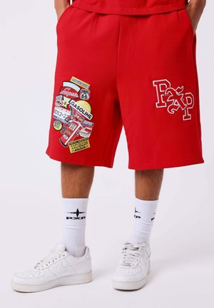 Persona con pantalones cortos rojos con parches coloridos de gasolina y de la Ruta 66, calcetines blancos y zapatillas blancas, de pie sobre un fondo claro.