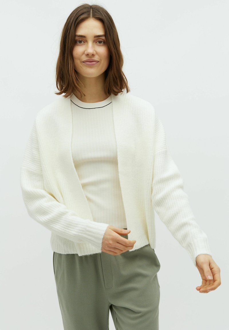 Vit stickad cardigan med öppen front, ribbat mönster och långa ärmar. Kombinerad med en figurnära krämfärgad topp och gröna byxor.