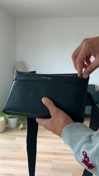 Geantă de umăr din piele neagră, cu formă rectangulară, având o închidere cu fermoar și curea ajustabilă. Logo-ul este afișat pe partea din față.