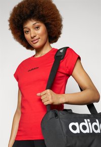 Crvena pamučna majica kratkih rukava s crnim "adidas" logom na prsima; model drži crnu torbu za sport s bijelim logom.