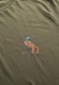 Olijfgroene katoenen t-shirt met een bedrukt ontwerp van een figuur in een oranje overall en blauwe helm, zittend met planten om zich heen.