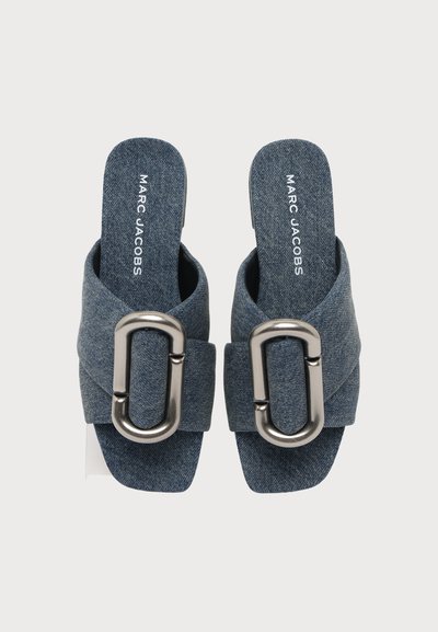 Ζευγάρι μπλε τζιν παντόφλες Marc Jacobs με μεγάλες ασημί αγκράφες στα λουράκια, σε θέα από ψηλά πάνω σε λευκό φόντο.