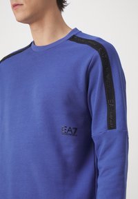 Uomo che indossa un felpa a maniche lunghe EA7 Emporio Armani blu con logo nero sul petto e strisce nere con marchio su spalle e maniche.