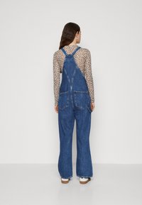 Temne modre jeans kombinezoni z ohlapnim krojem, nastavljivimi naramnicami in dvema žepoma na hrbtni strani. Nosi se jih čez bež dolgi rokav s potiskom leoparda.