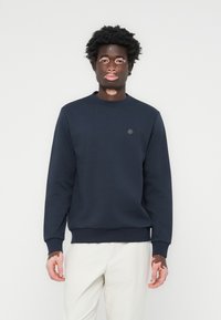 Marineblå sweatshirt laget av stoff, med rund hals, ribbestrikkede mansjetter og et lite merke på brystet. Glatt tekstur, avslappet passform.