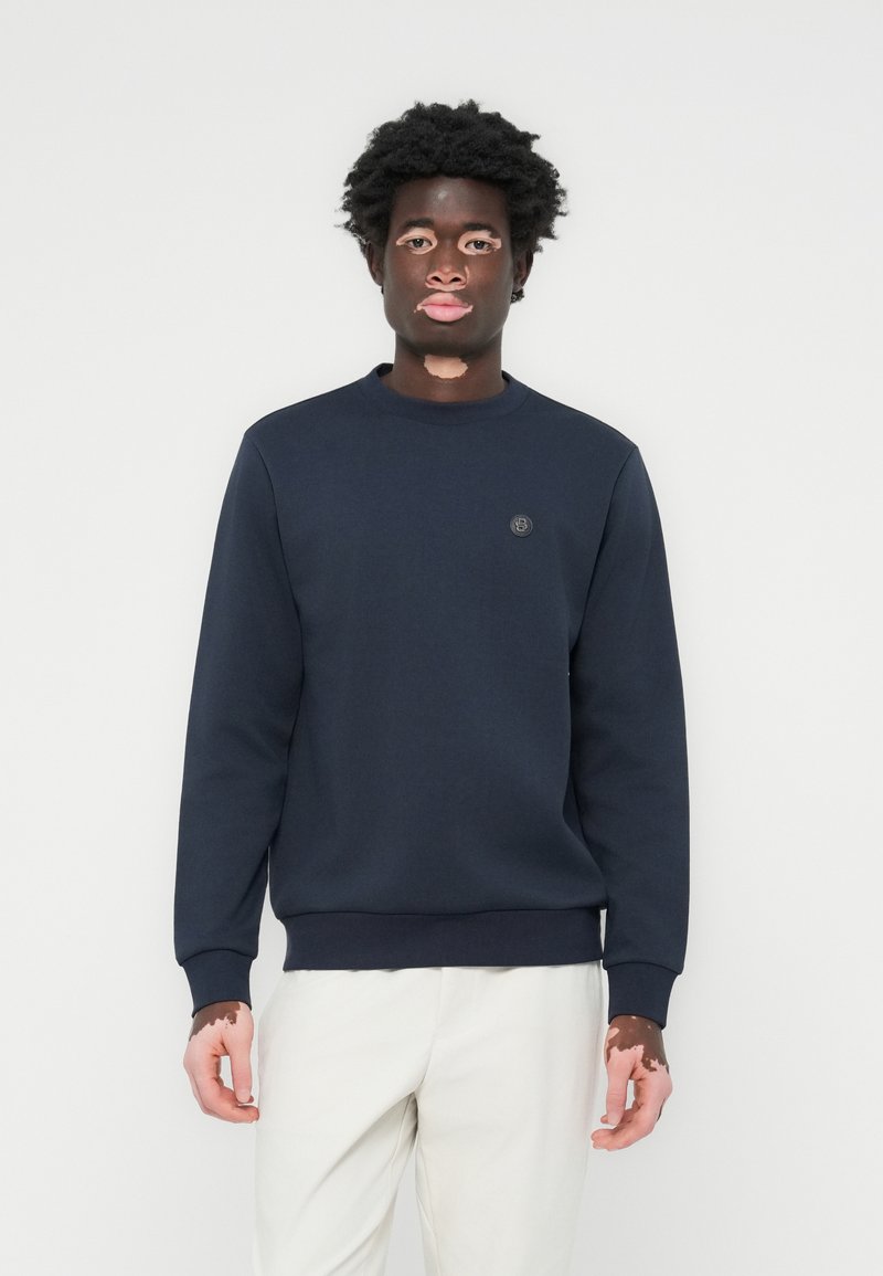 Marineblå sweatshirt laget av stoff, med rund hals, ribbestrikkede mansjetter og et lite merke på brystet. Glatt tekstur, avslappet passform.
