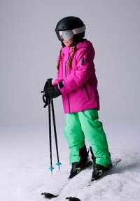 Vaikas ryškiai rožinėje striukėje ir žaliuose džinsuose stovi ant slidžių, laiko lazdelės, dėvi juodą šalmą ir akinius nuo saulės sniege.