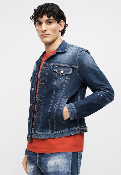 Jeune homme portant une veste et un pantalon en denim bleu avec des éclaboussures de peinture, sur un t-shirt rouge, debout avec une main dans la poche de la veste.