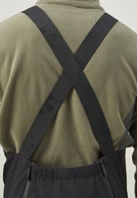 Sorte justerbare stropper krydser over en olivengrøn fleece. Stroppene har et logomønster og er fastgjort til et mørkt yderstof i taljen.