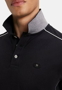 Polo noir avec un col gris, texture côtelée, patte de boutonnage à trois boutons, coutures blanches accentuées et un logo rond sur la poitrine.