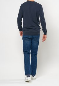 Man som bär en marinblå tröja, blå jeans och vita sneakers går bort från en ljus, enfärgad bakgrund.