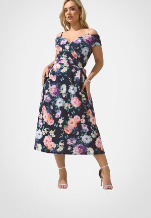 Kvinde iført en navyblå off-shoulder midi-kjole med flerfarvet blomsterprint og sølvfarvede højhælede sandaler, poserede med hænderne i lommerne.