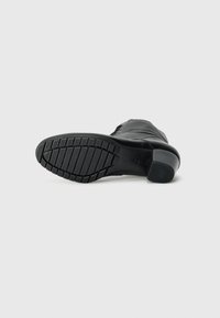 Bottine en cuir noir avec une semelle en caoutchouc, présentant un motif de semelle texturé et un talon carré. Design minimaliste sans embellissements visibles.