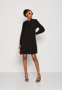 Svart lagerdress med långa ärmar och knäppning, accentuerad med en rosa handväska. Modellen bär stora solglasögon och klackar med remmar.