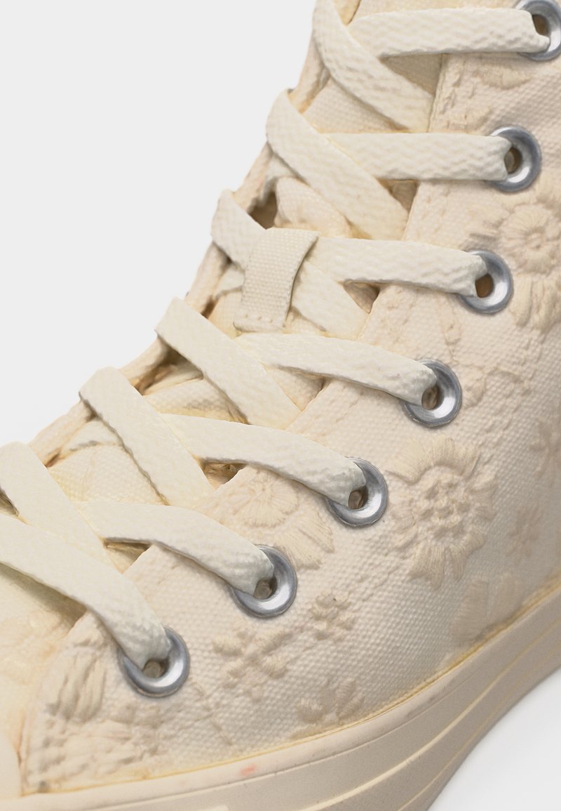 Cremefarbener High-Top-Sneaker mit stoffartiger Blumenstickerei und dicken weißen Schnürsenkeln, die durch Metallösen geführt sind.
