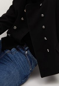 Czarny dwurzędowy płaszcz z srebrnymi guzikami, zestawiony z niebieskimi dżinsami. Płaszcz charakteryzuje się przemyślaną konstrukcją i gładką fakturą tkaniny.