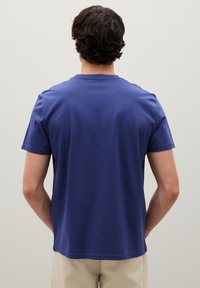 Mann von hinten zu sehen, der ein schlichtes blaues T-Shirt mit kurzen Ärmeln und beige Hosen trägt, vor einer einfarbigen hellgrauen Wand.