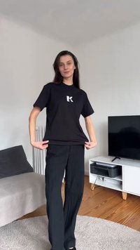 Νέα γυναίκα με μαύρο παντελόνι και μαύρο μπλουζάκι με λευκό λογότυπο «K» στέκεται σε ένα σαλόνι με τηλεόραση, καναπέ και ξύλινο πάτωμα.