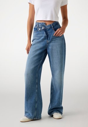 Light wash denim wide-leg jeans with a high waist, featuring dual buttons, and a relaxed fit down to the hem, paired with white sneakers.