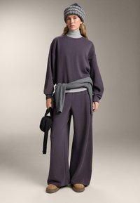 Felpa viola oversize e pantaloni a gamba larga, abbinati a un maglione grigio annodato in vita e un cappello di maglia grigio a fantasia. Calzature marroni.