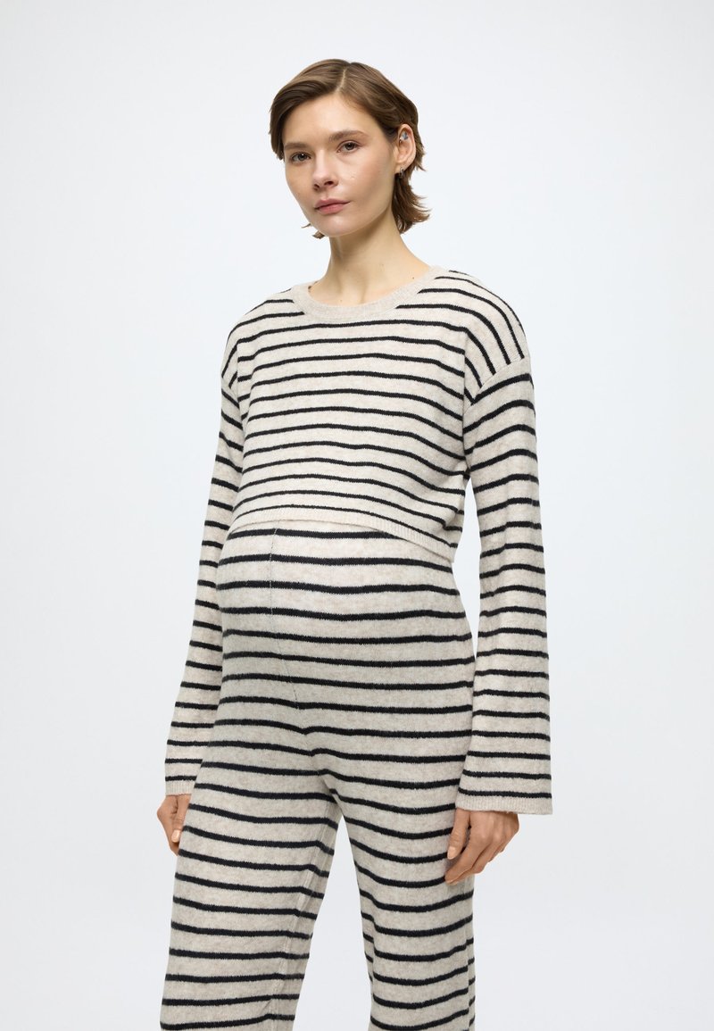 Csíkos kötött kismama ruha világos bézs színben fekete csíkokkal. Laza szabású felsővel és bőszárú nadrággal, kiemelve a kerek hasat.