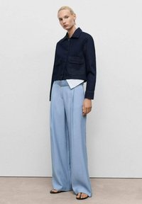 Dunkelblaue, kurze Jeansjacke mit zwei vorderen Taschen, über einem weißen Hemd mit Kragen getragen, kombiniert mit hellblauen, weit geschnittenen Hosen und schwarzen Sandalen.