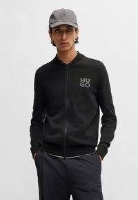 Sudadera negra con cremallera y cuello redondo, con el logo "HUGO". El material parece suave y de punto, con un diseño ajustado y puños acanalados.
