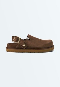 Bruine suède klomp met verstelbare gesp en dikke rubberen zool, ontworpen voor slip-on comfort en casual gebruik.