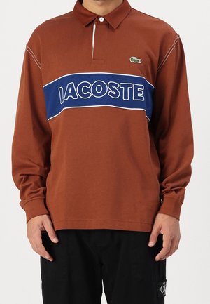 Brązowa koszulka polo z długim rękawem, z niebiesko-białym poziomym paskiem i napisem „LACOSTE”. Materiał jest gładki, z klasycznym kołnierzykiem i subtelnym logo.