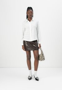 Camicia bianca a maniche lunghe con motivo ad occhiello, abbinata a una mini gonna lucida in pelle di coccodrillo marrone. Tieni in mano una borsa in pelle di serpente. Scarpe nere e calze bianche.