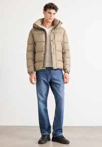 Doudoune beige avec capuche, dotée d'une fermeture éclair et de boutons-pression, associée à un jean en denim bleu et à des baskets noires.