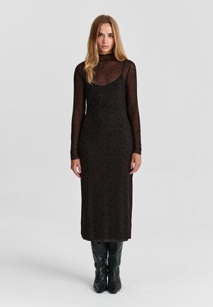 Femme aux longs cheveux blonds portant un haut noir transparent à manches longues sous une robe noire à imprimé léopard et des bottes en cuir noires, debout sur un fond gris.