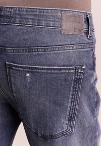 Vaqueros de mezclilla azul con un acabado desgastado, presentan una etiqueta de parche en la cinturilla y dos bolsillos traseros con detalles de costura visibles.