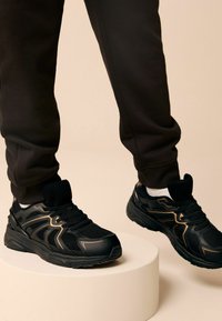 Chaussures de sport noires en matériaux en mesh et en caoutchouc, avec des accents dorés, une surface texturée et un bout arrondi. Portées avec un pantalon noir.