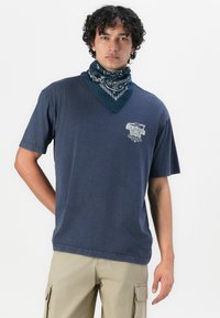 Marinblå kortärmad t-shirt med v-ringning och grafiskt tryck. I kombination med en teal bandana med vita mönster runt halsen.