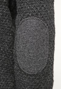 Donkergrijze gebreide trui stof met een gestructureerd oppervlak, voorzien van een lichtergrijze vilten elleboogpatch en een close-up van de stiksels.